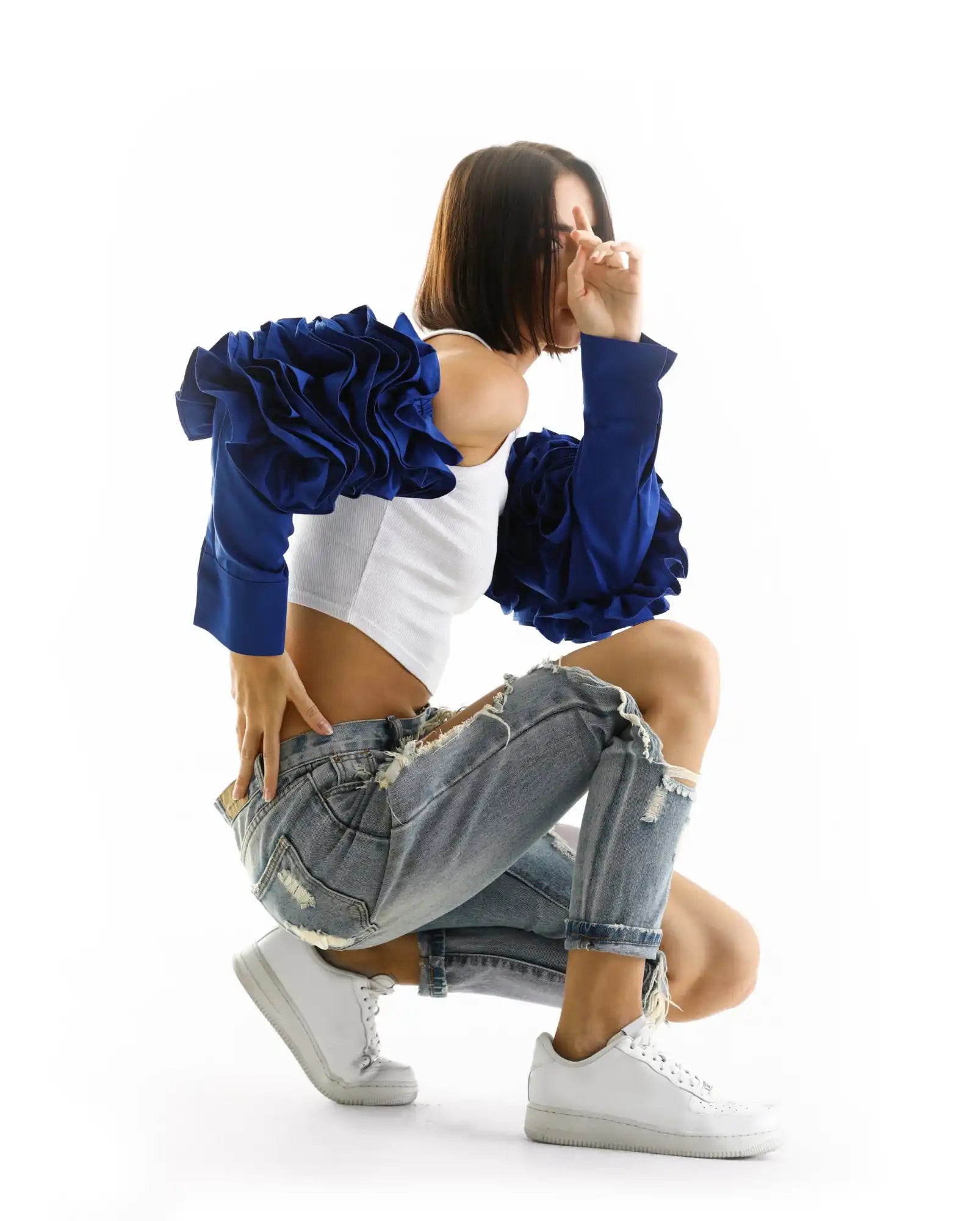 A dancer in ripped jeans, white crop top and blue ruffled jacket crouches in a stylized pose wearing white sneakers.