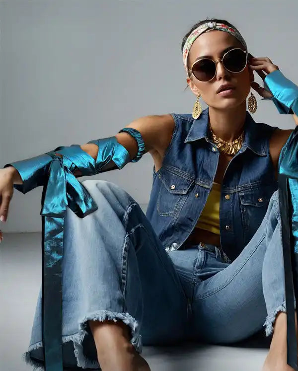 A fashion model wearing a denim vest, metallic blue sleeves, sunglasses, and gold jewelry while sitting in ripped jeans.