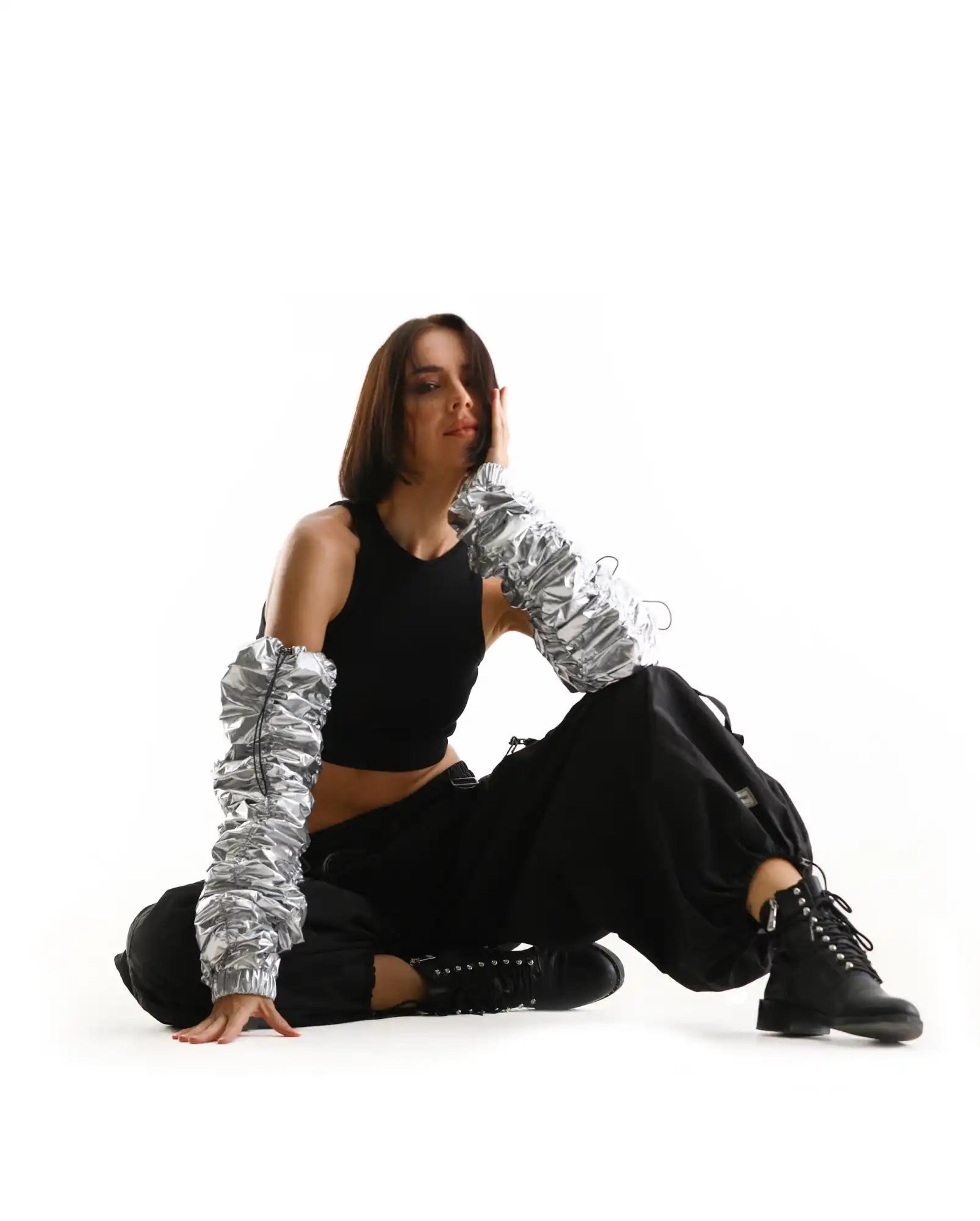 A person wearing a black crop top, cargo pants, combat boots and metallic silver puff sleeves sits on the ground in a relaxed pose.